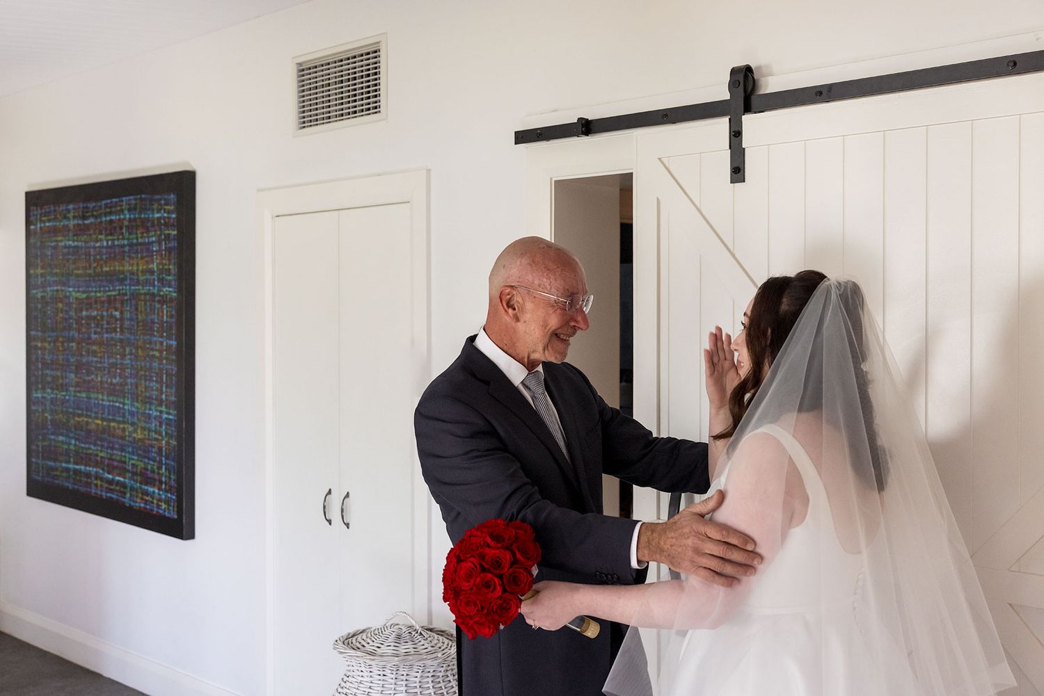 A bride in a white veil and dress stands with her Dad in a dark suit, surrounded by white walls, seeing her for the first time in her dress.