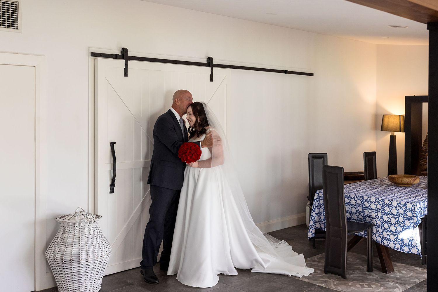 A bride in a white veil and dress stands with her Dad in a dark suit, surrounded by white walls, seeing her for the first time in her dress.