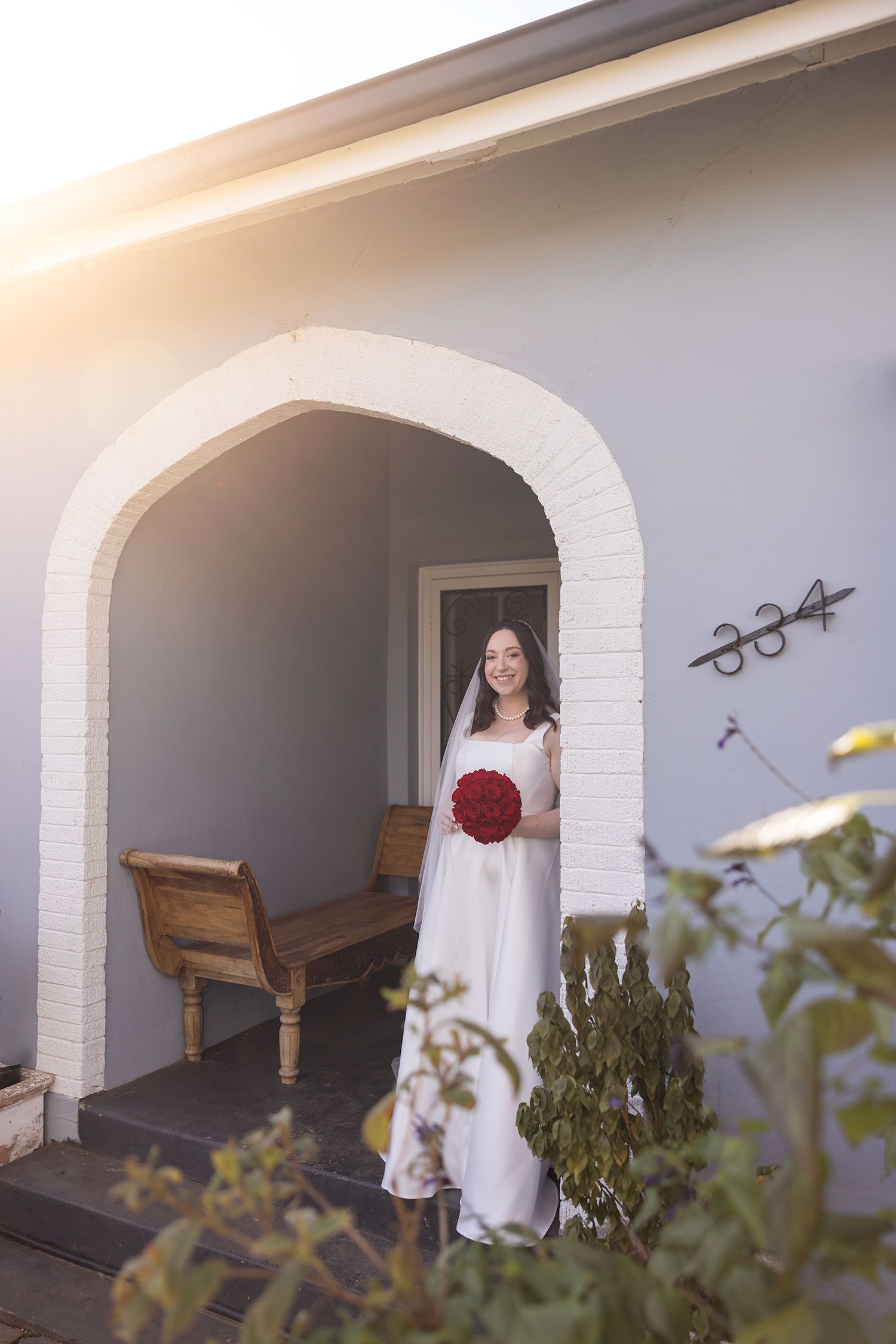 A Bride wearing her Anna Campbell wedding dress holding red roses stands in the old arch entrance of an Albury house soft sunlight.