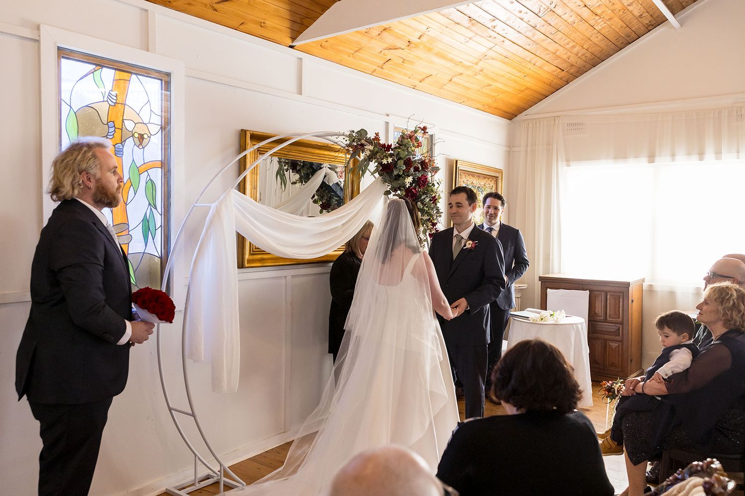Wedding guests gathered in a bright cottage interior with wood-paneled ceiling and white fabric decorations at La Maison Wodonga.