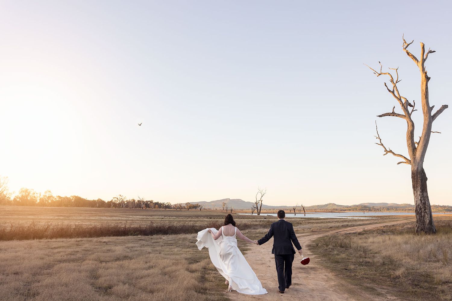 Two people walking hand in hand along a scenic country landscape at sunset with bare trees and the Hume Weir in the background.