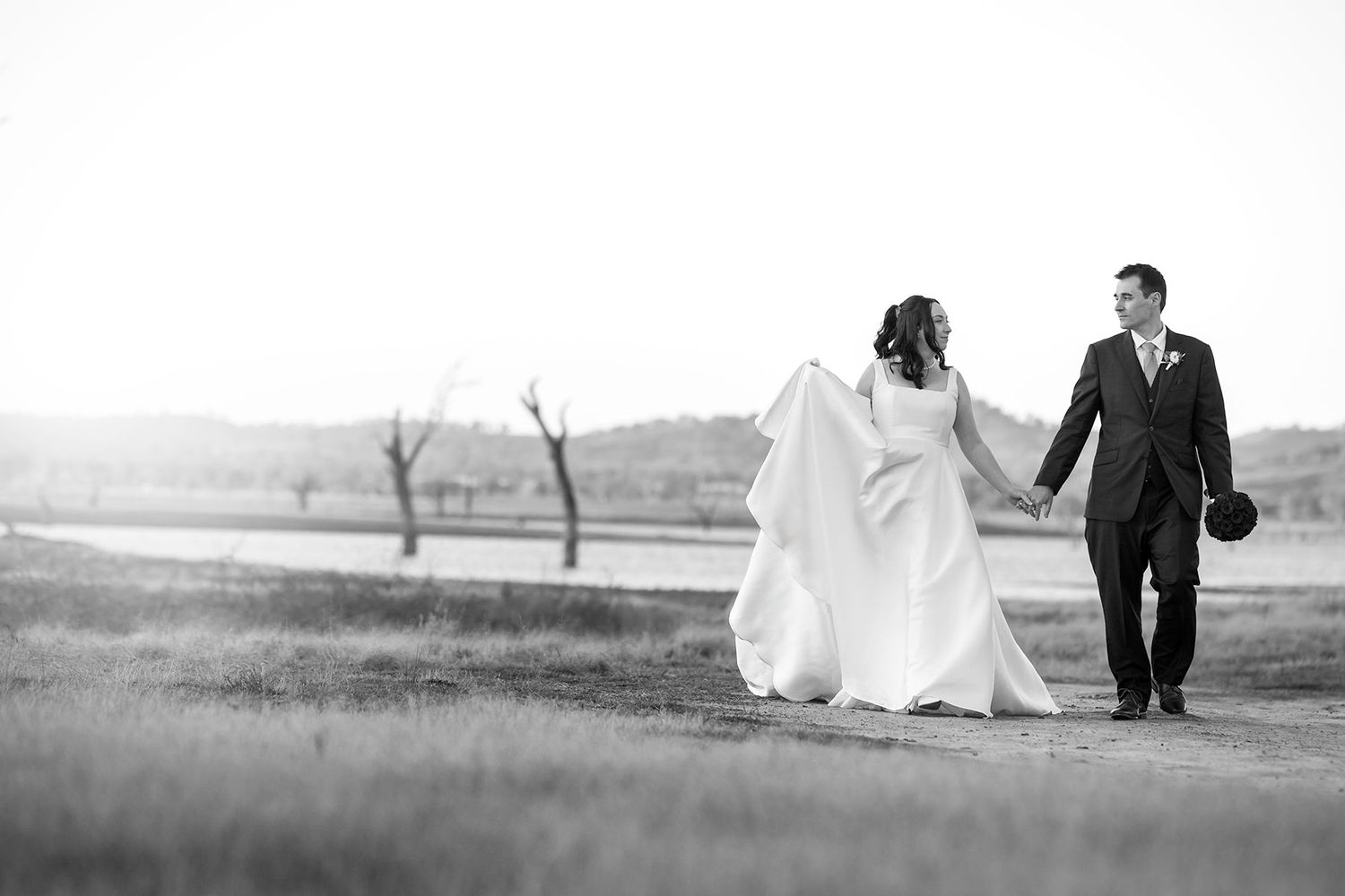A Bride and Groom walking hand in hand along a scenic country landscape at sunset with bare trees and the Hume Weir in the background.