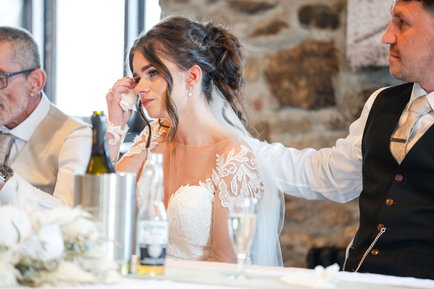 Wedding reception guests raise glasses in a toast at rustic stone venue with wood beams and natural light.