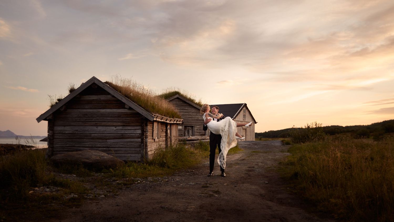 Groom carrying his bride close to old, traditional wooden houses (laftahus) with pastel skies and midnight sun