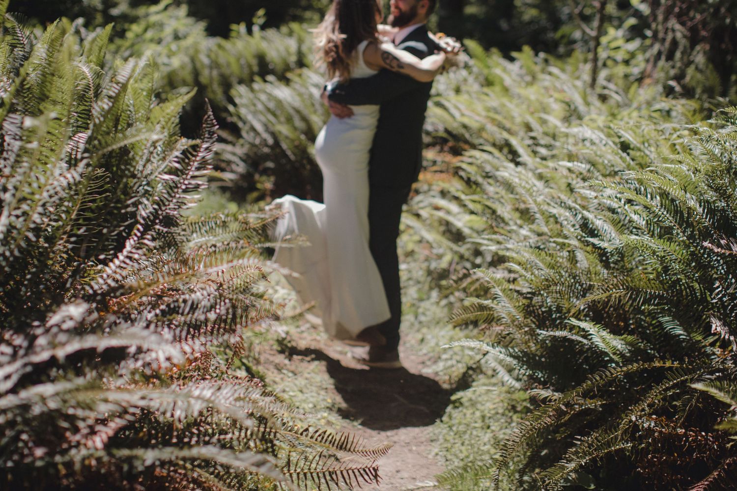 A person walks through a lush garden path surrounded by green ferns and foliage in dappled sunlight.