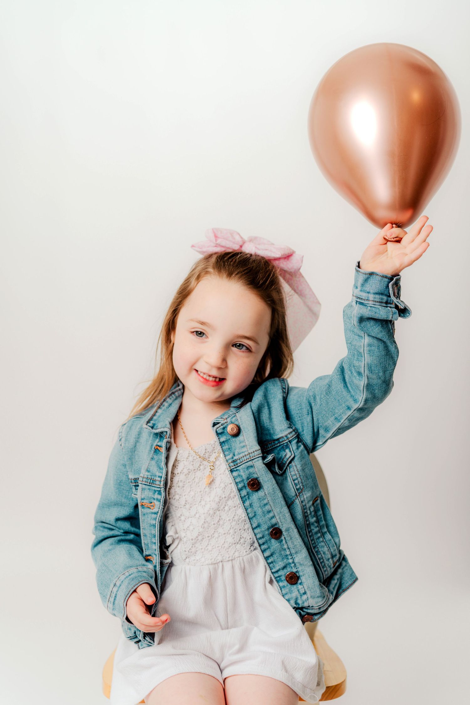 A series of playful poses with a child wearing a denim jacket and white dress holding rose gold balloons against a white backdrop.