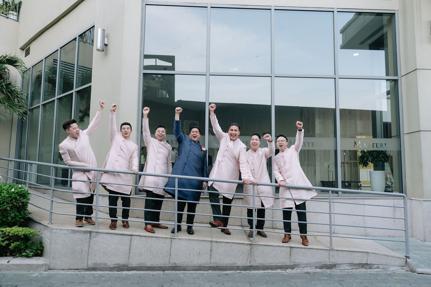 Groom and groomsmen in ao dai celebrating joyfully with raised fists outside a modern building.