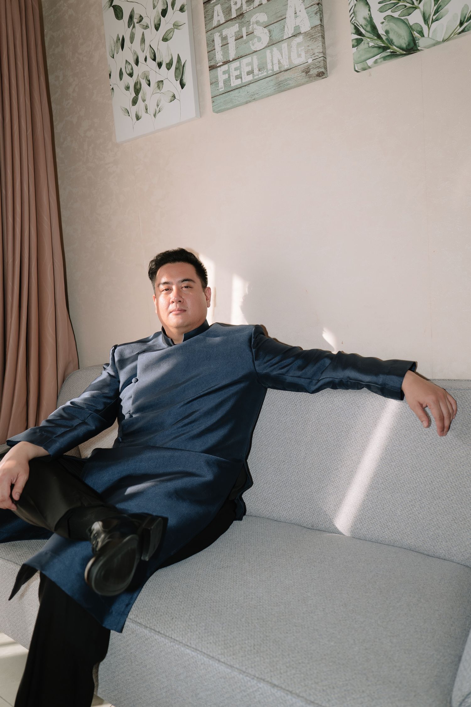 Groom in navy blue ao dai relaxing confidently on a gray sofa in a sunlit room.