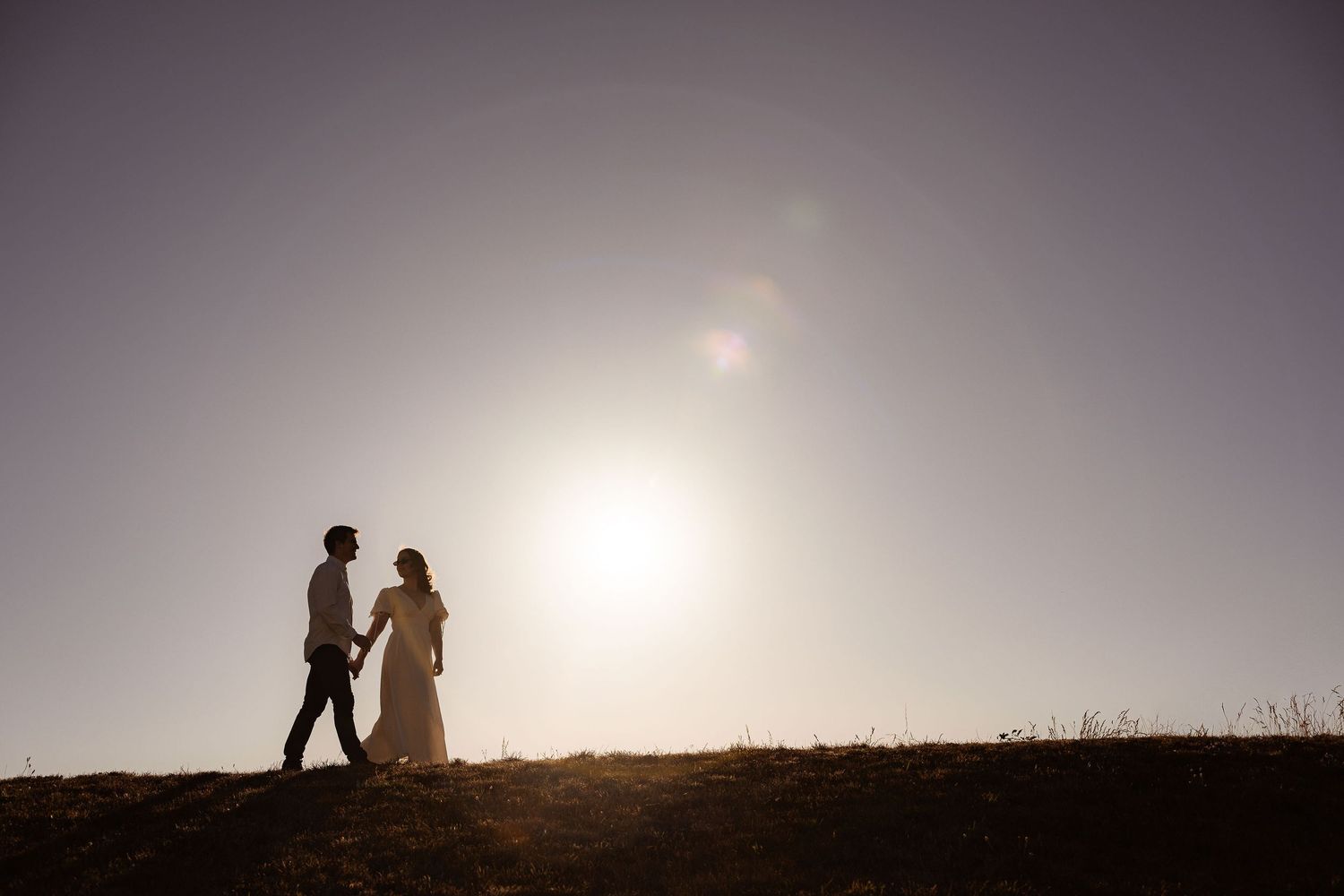 A sequence of silhouetted figures sit together on a hill against a bright sunset horizon.