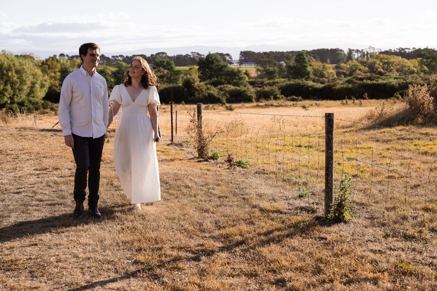 Couple in white and white attire share an intimate moment in golden sun-dried grassland at sunset.