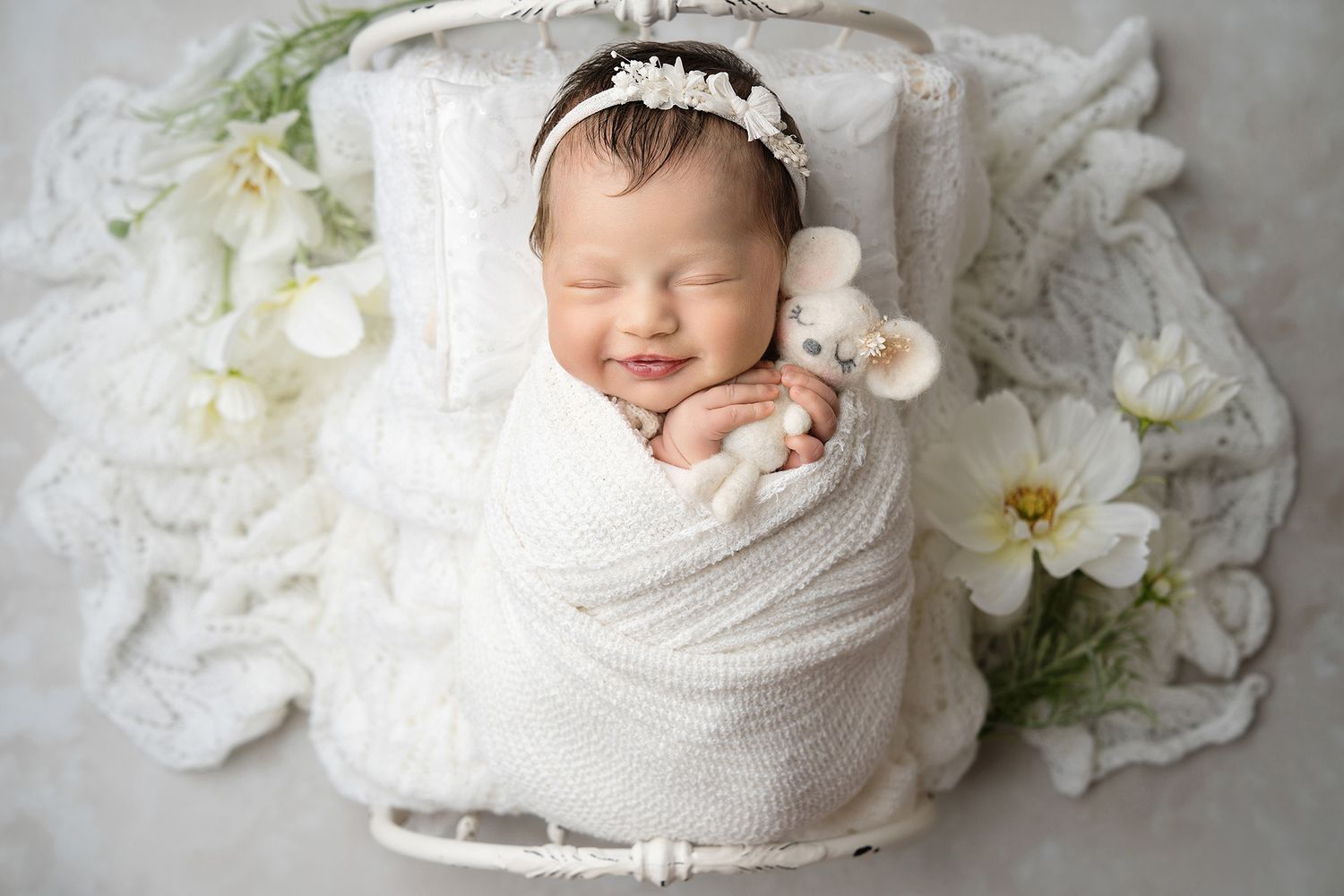 Newborn baby wrapped in white with lace headband surrounded by white flowers.