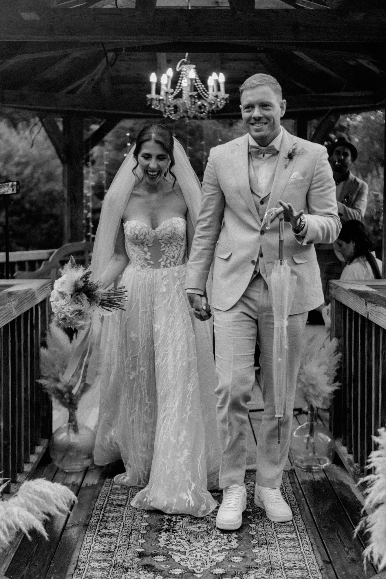 A wedding couple walks down a decorated rustic bridge under a chandelier.