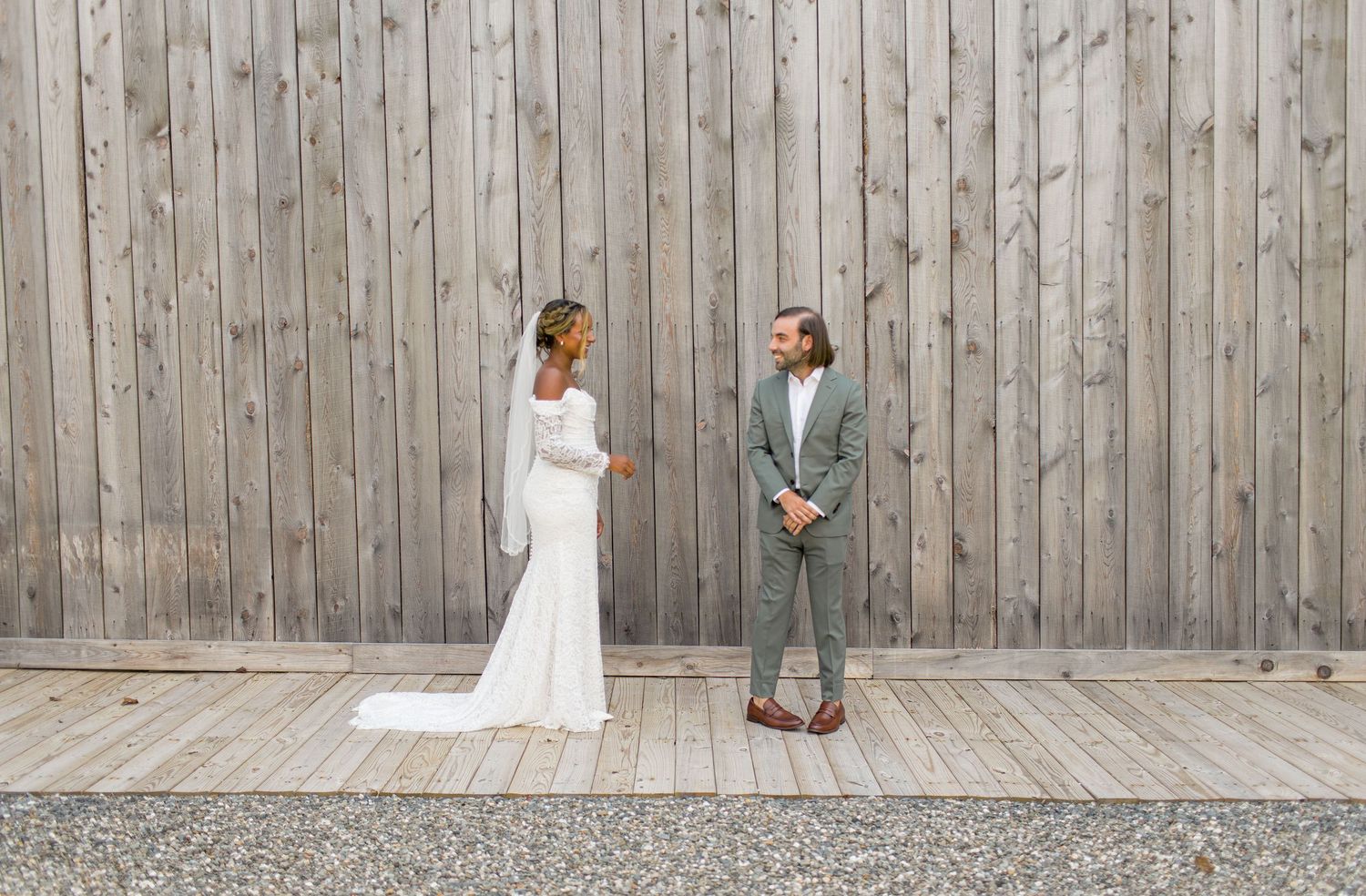 A couple in wedding attire stands apart against a rustic wooden wall during a modern minimalist outdoor photoshoot.