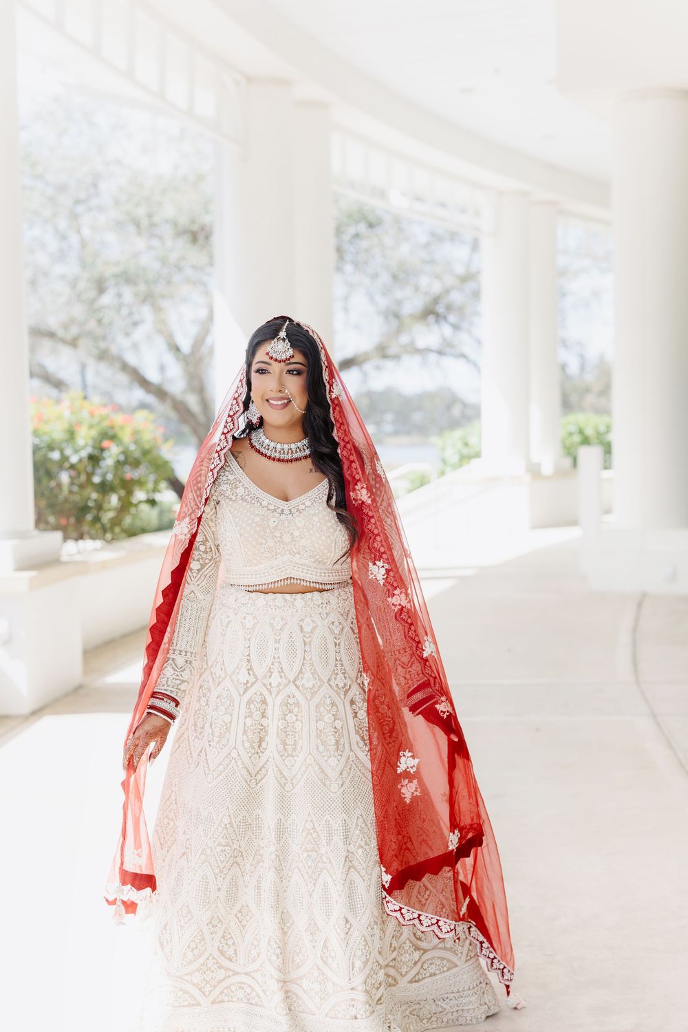 A bride in a white embroidered dress and red dupatta poses against a white colonnade backdrop.