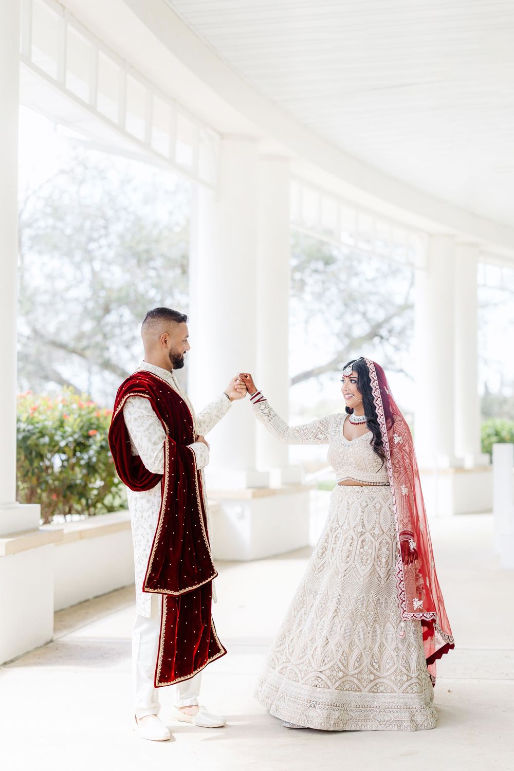 A couple in traditional Indian wedding attire pose in a white-columned corridor for their bridal photoshoot.