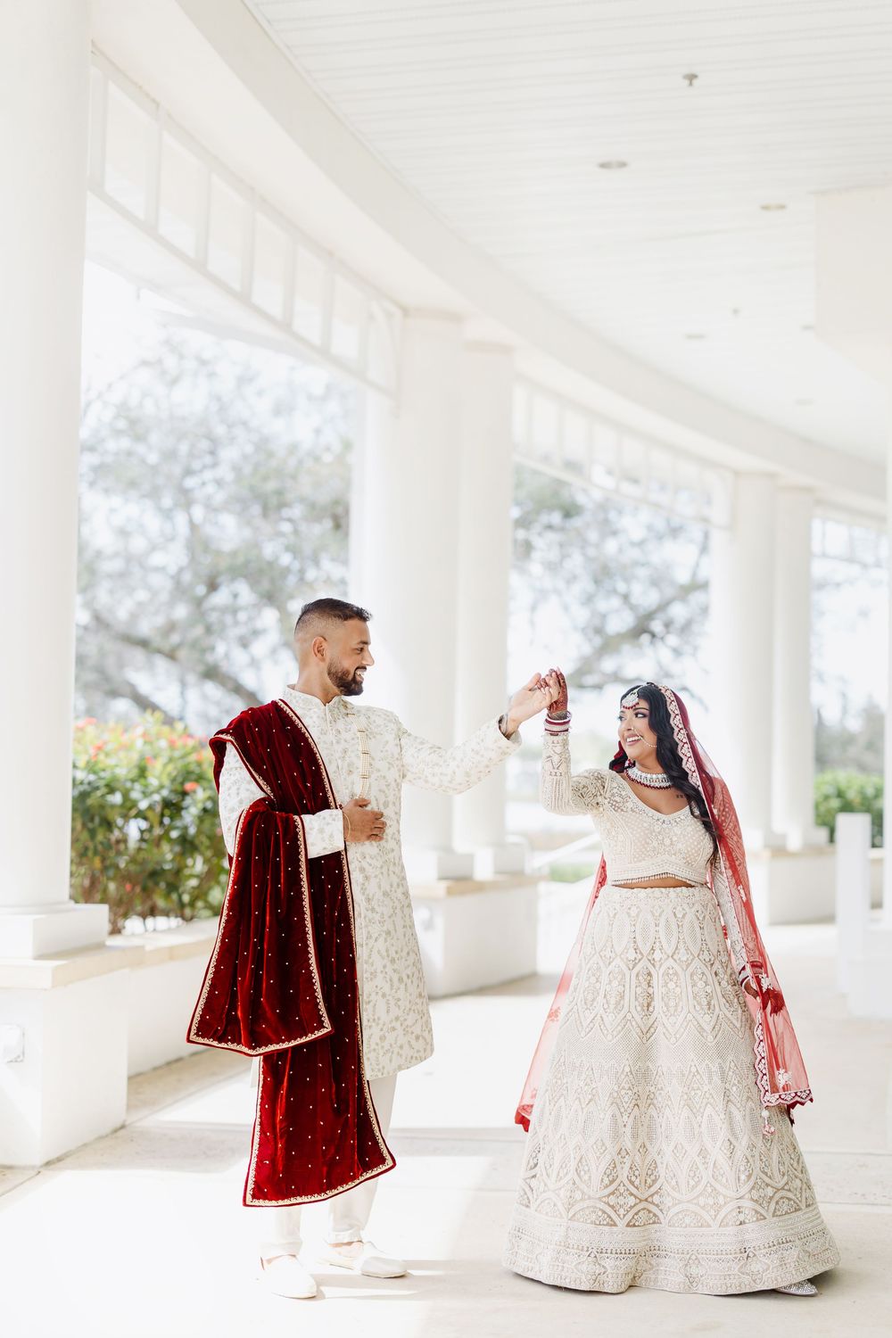 A couple in opulent Indian wedding attire poses in white pillared venue during their ceremony photo session.