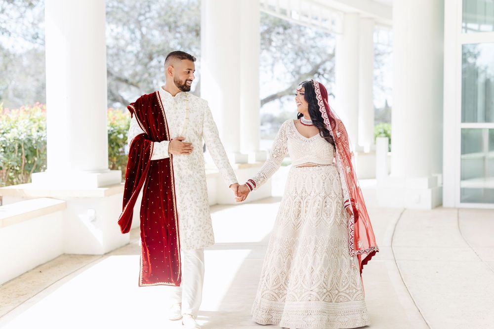 A couple in traditional South Asian wedding attire holds hands under an elegant white columned porch.