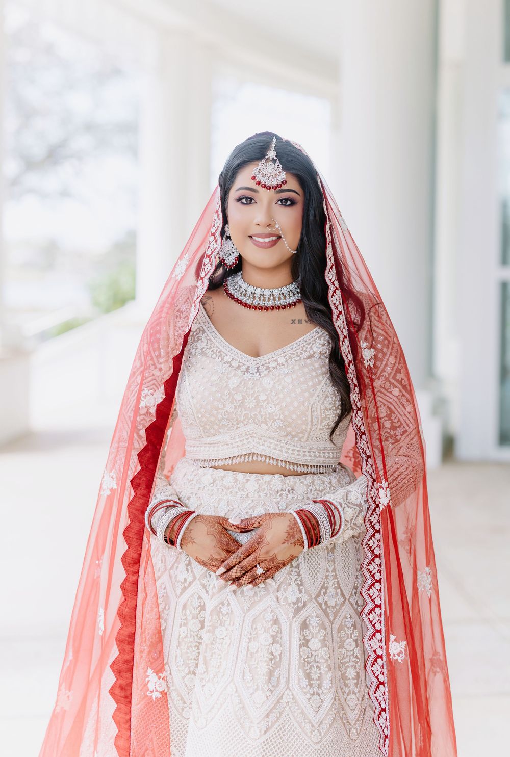 South Asian bride in white and silver lehenga with coral dupatta and traditional wedding jewelry stands elegantly.