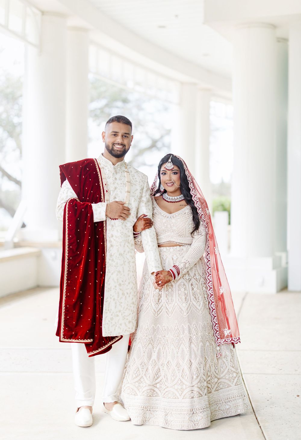 A couple in traditional South Asian wedding attire, with white and red garments, pose together in an elegant indoor setting.