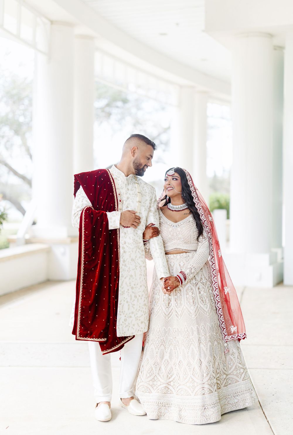 A couple in traditional South Asian wedding attire share a sweet moment in white colonnade, wearing ornate red and white outfits.