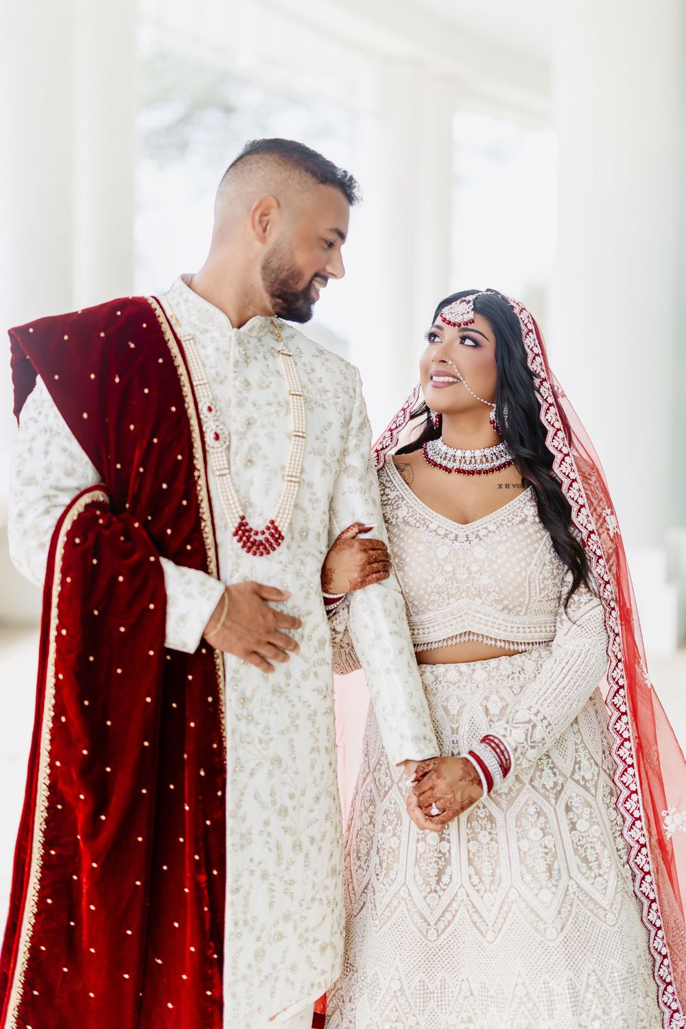 Newlyweds in traditional Indian wedding lehenga and sherwani share joyful moment under white pillars.