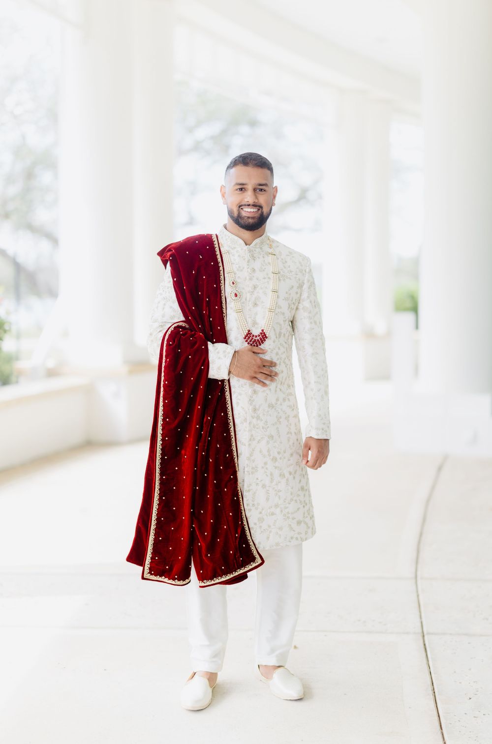 South Asian groom poses in white embroidered sherwani with red velvet dupatta at wedding photoshoot.