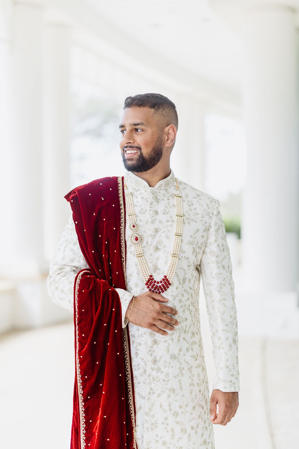 A wedding attire featuring an elegant white embroidered sherwani and deep red velvet dupatta with sparkly details.