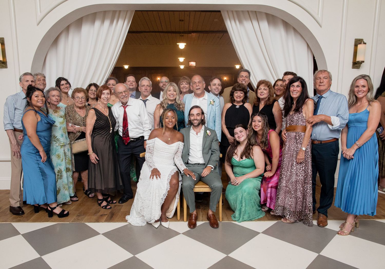 A group of wedding guests posing together for a panoramic photo in a reception venue with white walls.