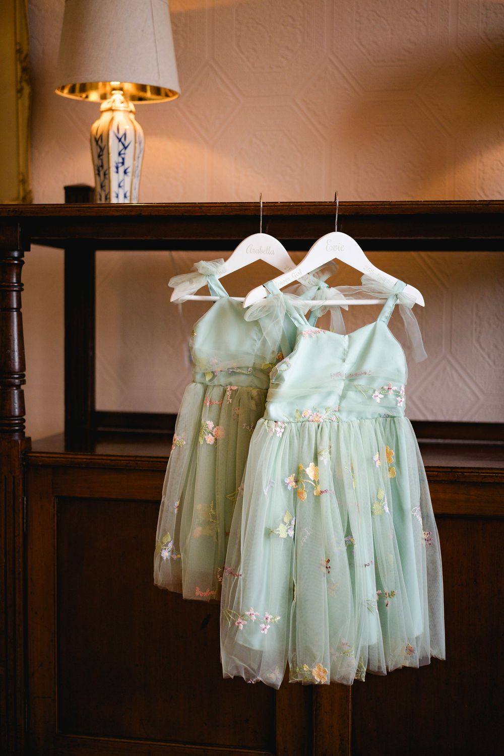 Delicate mint green flower girl dresses with floral details hang on white hangers in a row.