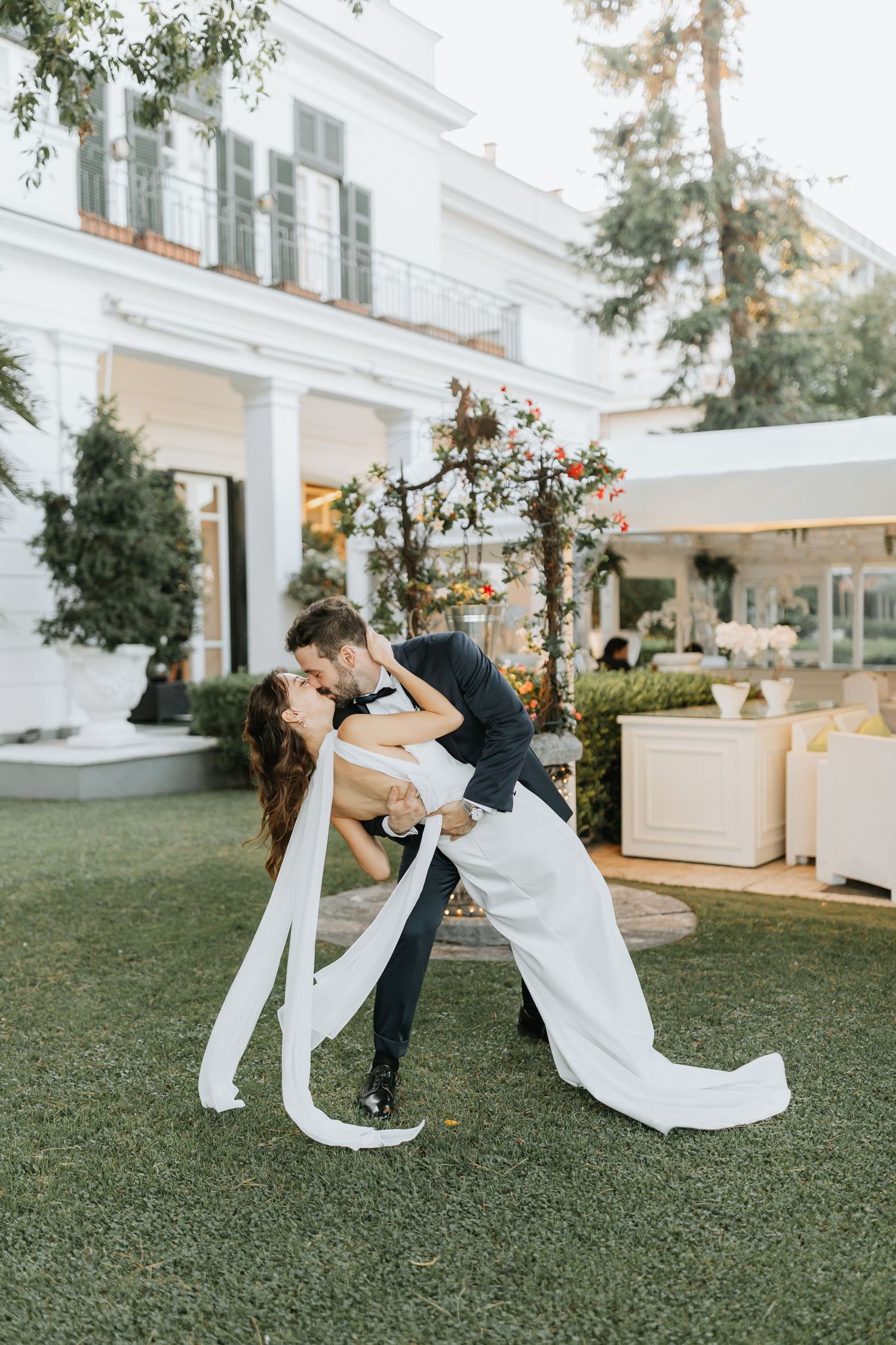 Romantic couple dancing and dipping on green lawn in front of elegant white mansion at sunset. Destination Wedding Photographer, Italy