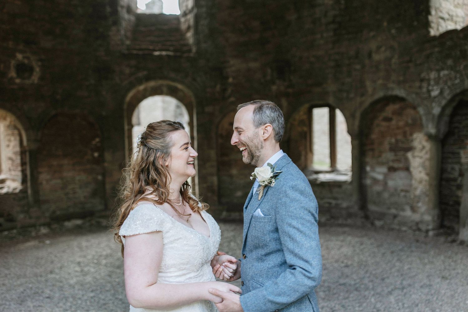 A romantic wedding couple stands together in front of weathered brick walls at their outdoor celebration.