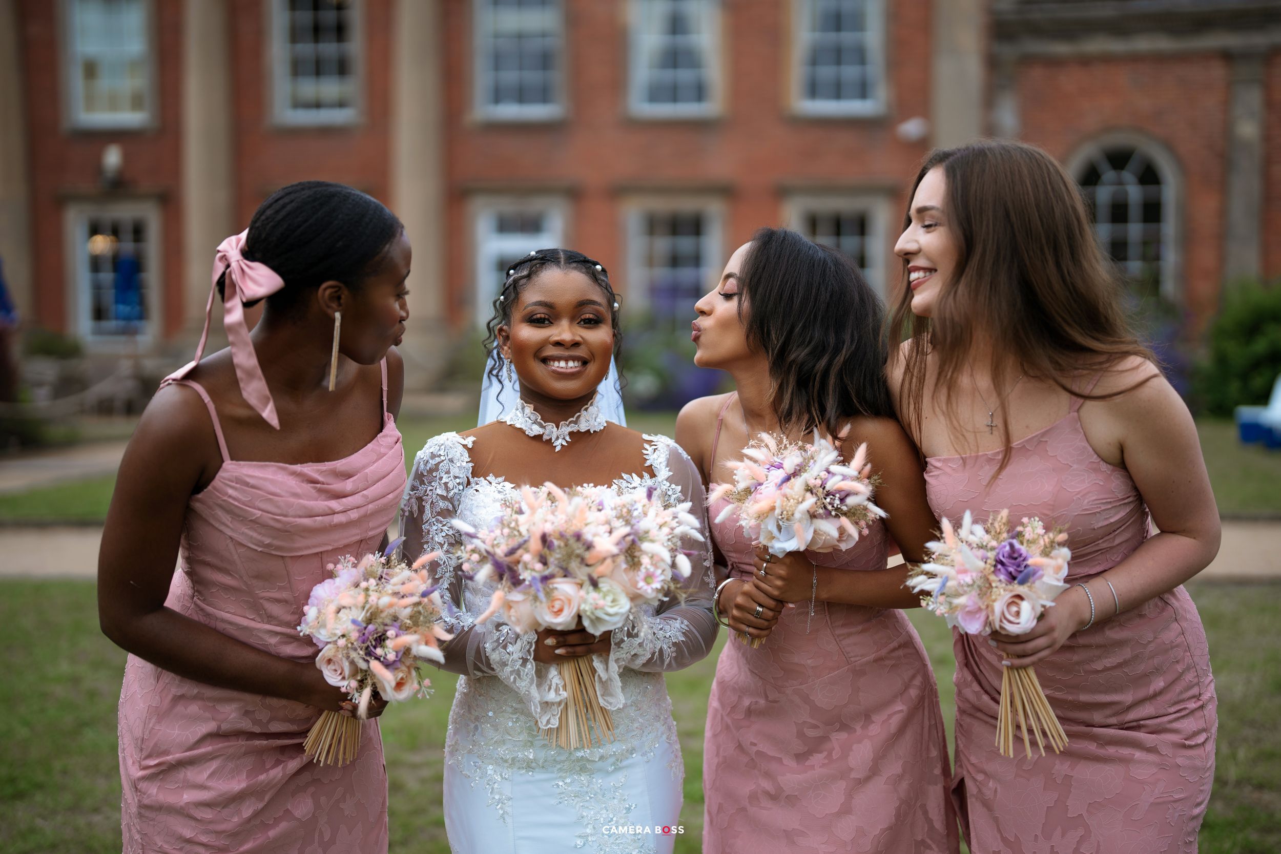 A group of wedding attendants in pink dresses laugh together while holding bouquets in a formal garden setting.