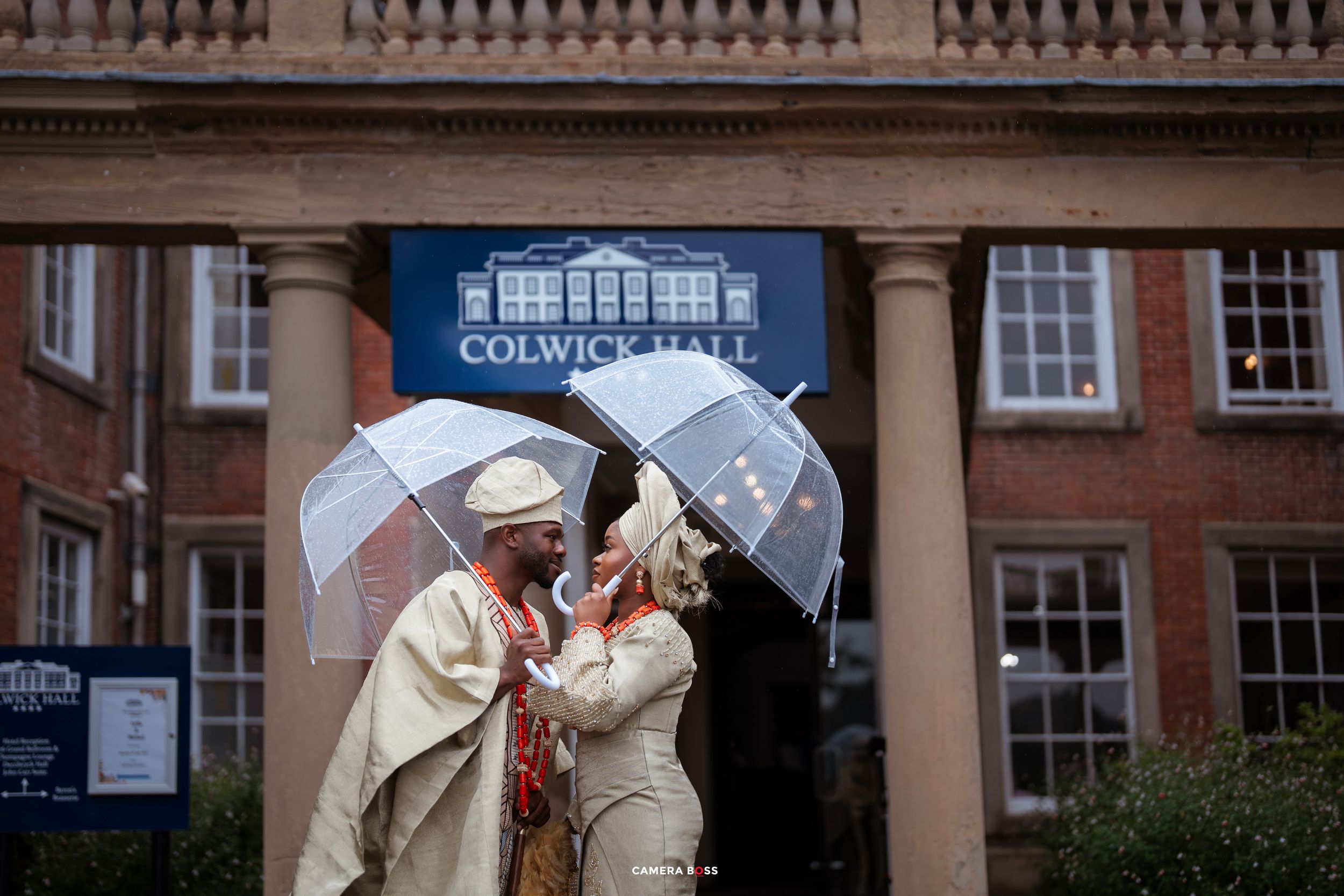 A wedding couple in white attire share a romantic kiss under umbrellas in front of a historic brick building.