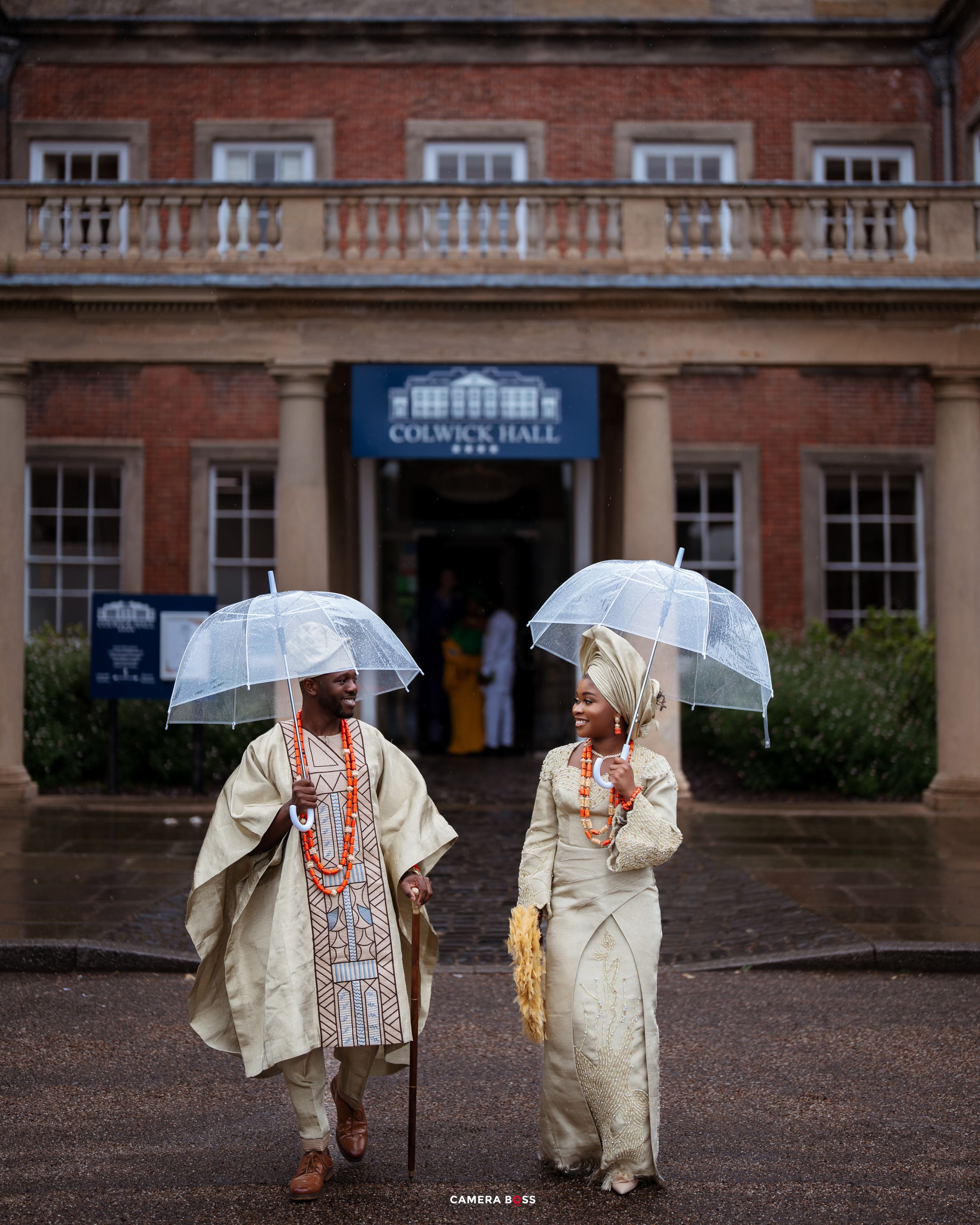 People in light colored clothing holding clear umbrellas stand on a wet brick courtyard outside a historic building.