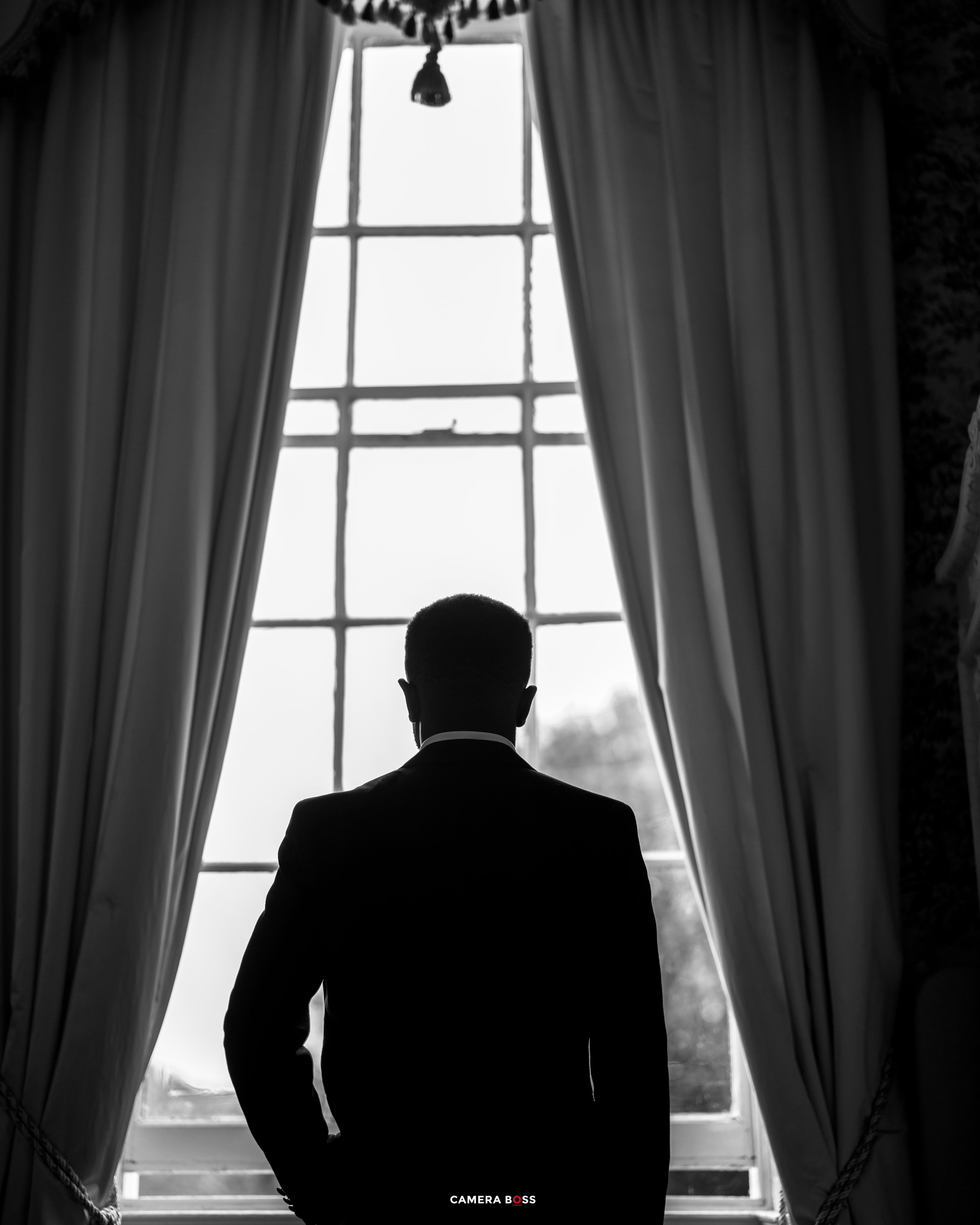 A dramatic silhouette of someone standing pensively by a window with long curtains in black and white.