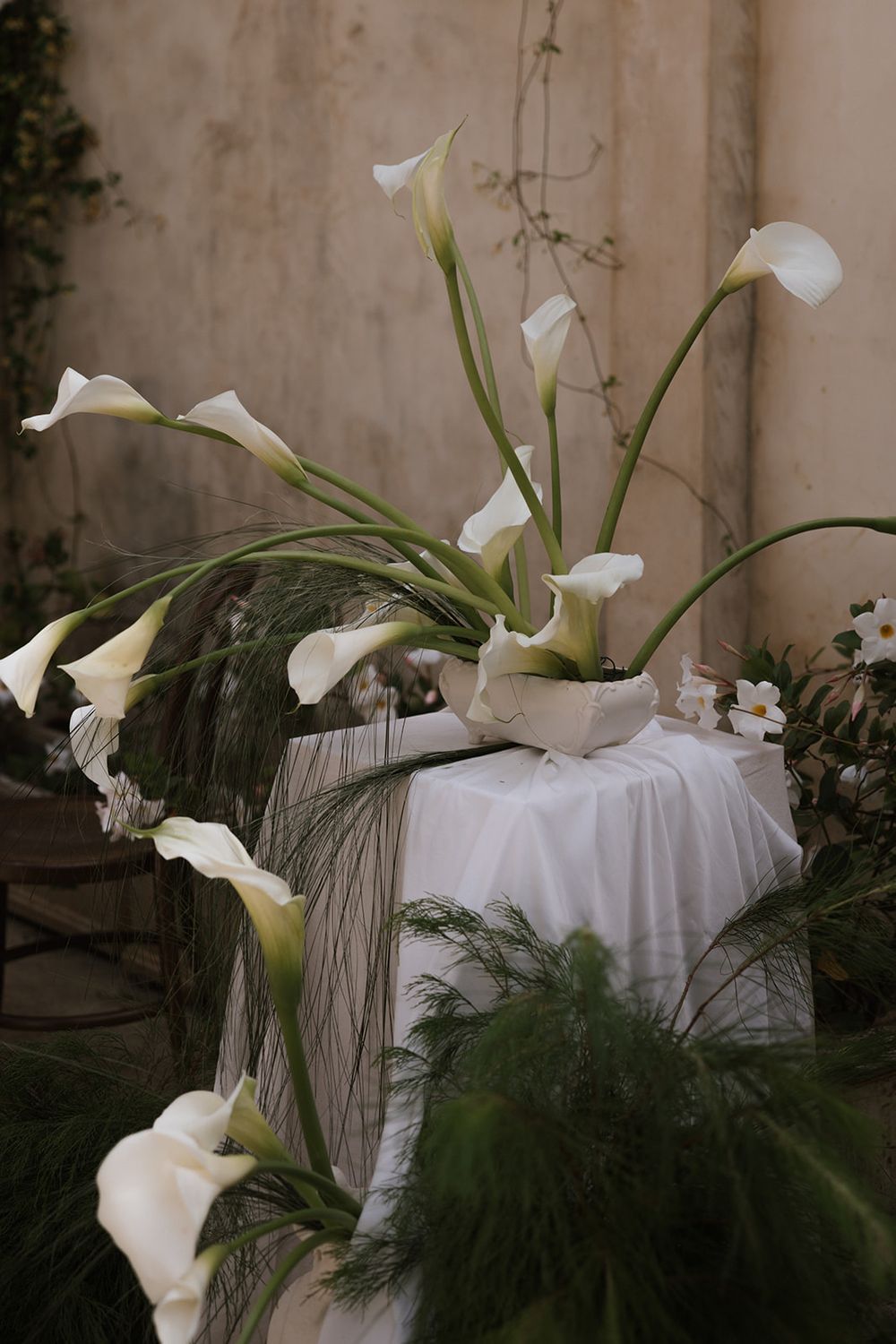 Elegant white calla lilies and delicate greenery arranged on white linens create a serene botanical display.
