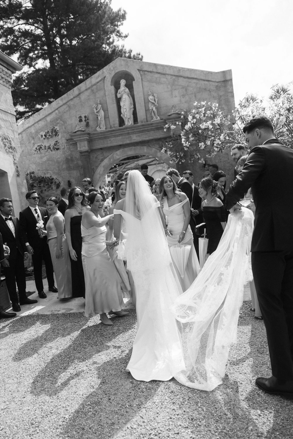 Wedding party in formal attire gathers outdoors on a gravel path during an elegant celebration.