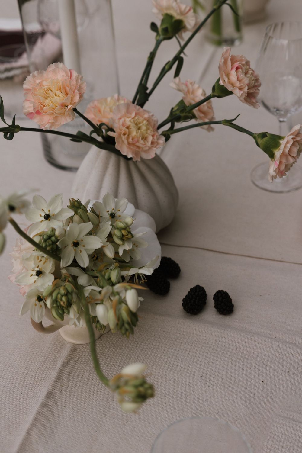 Delicate pink carnations and white flowers arranged in a gray vase with blackberries on a light table setting.