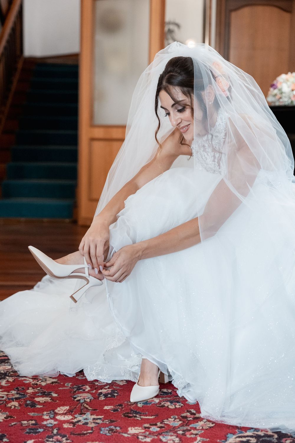A bride in a white wedding dress and veil adjusts her gown while sitting on an ornate red patterned carpet.