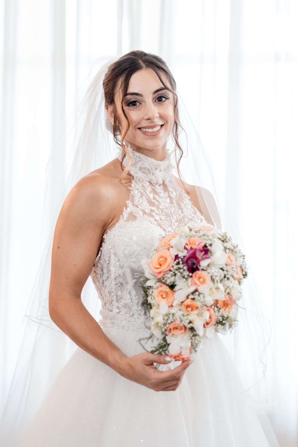 Beautiful bridal portraits showcasing an elegant white lace wedding dress and peachy pink floral bouquet against white curtains.