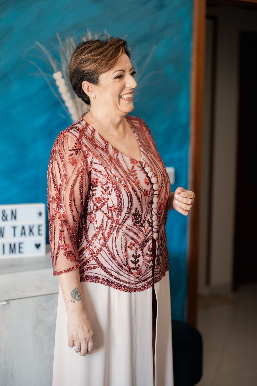 Wedding guest wearing a sequined pink top with white dress poses in front of a blue backdrop during celebration.