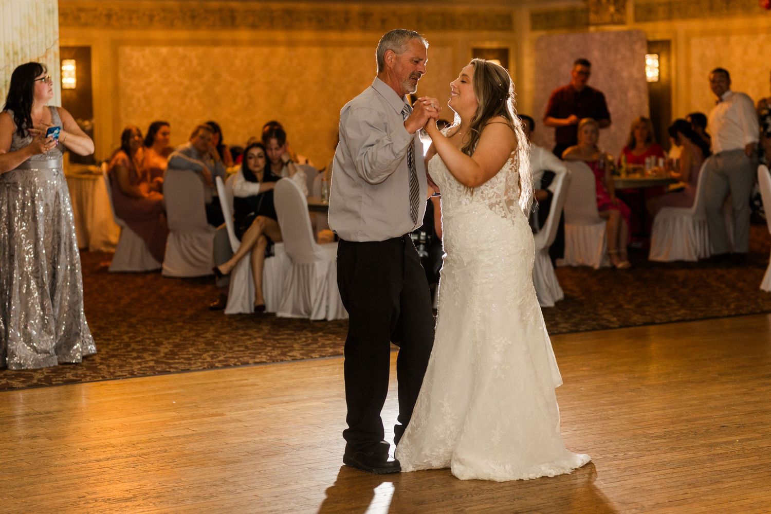 A couple shares their first dance at a wedding reception in a warmly lit ballroom with guests in the background.