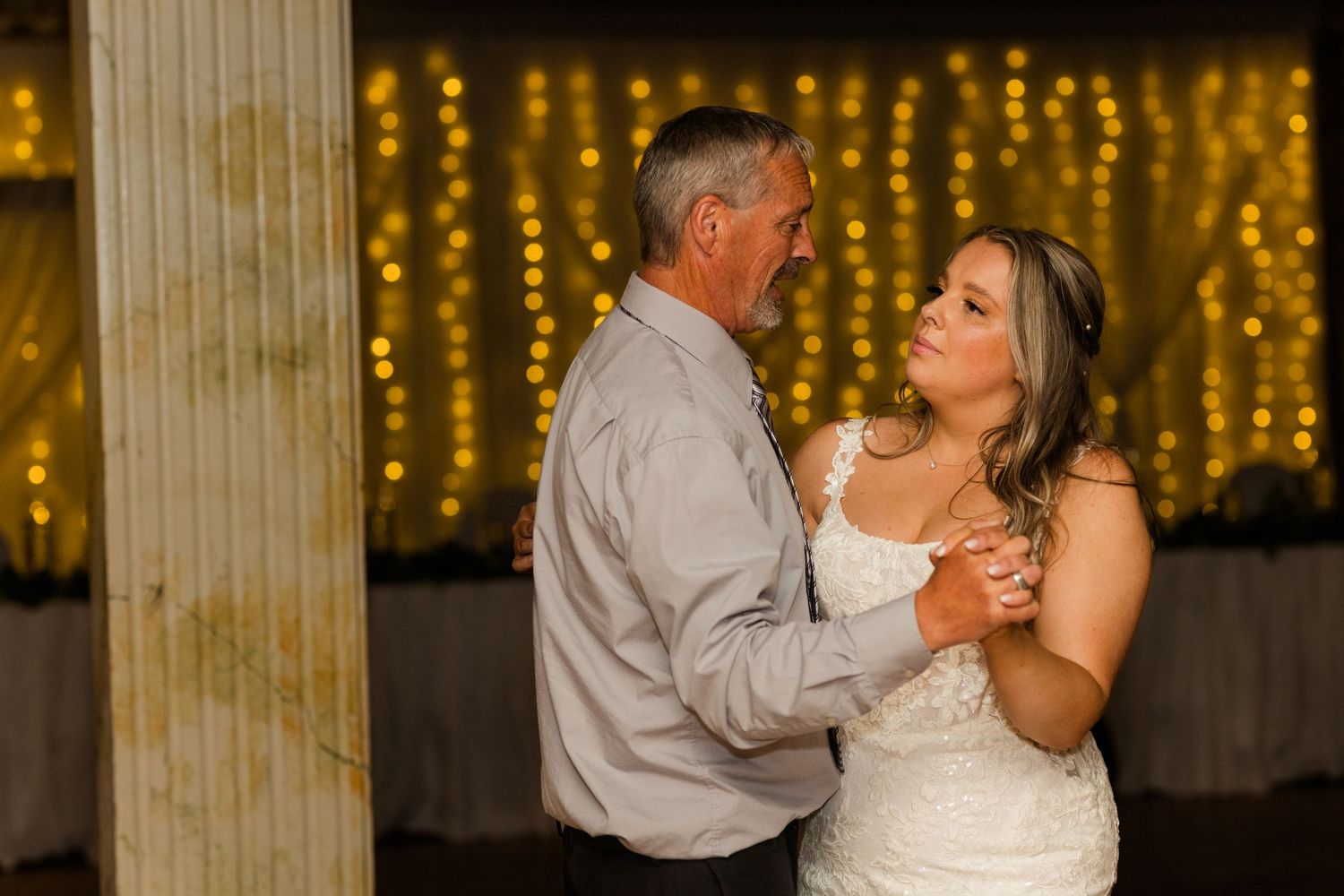 A couple wearing formal attire shares a romantic first dance at a wedding reception with glowing lights in the background.