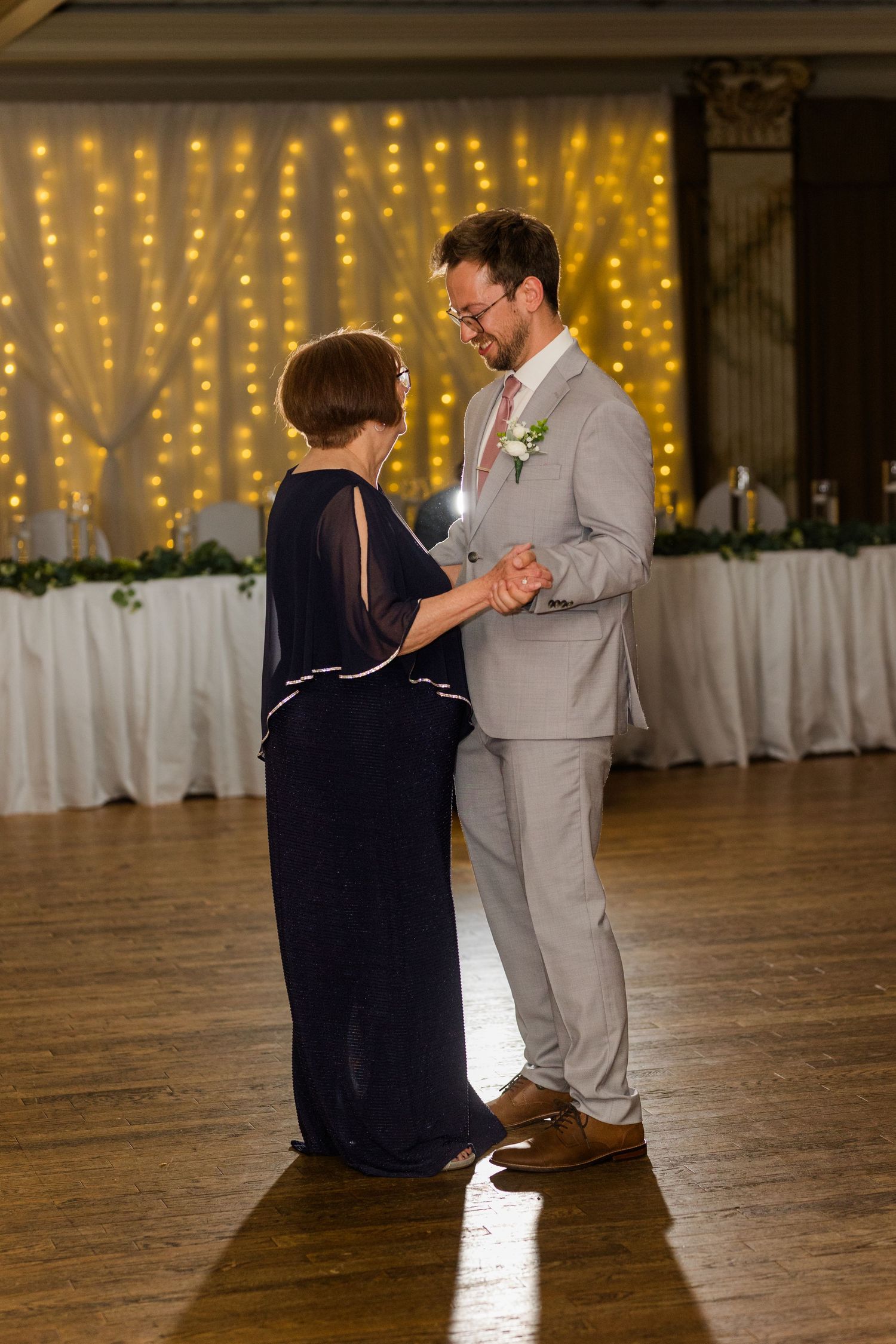 Two dancers practice their steps on a wooden dance floor with twinkling string lights glowing in the background.