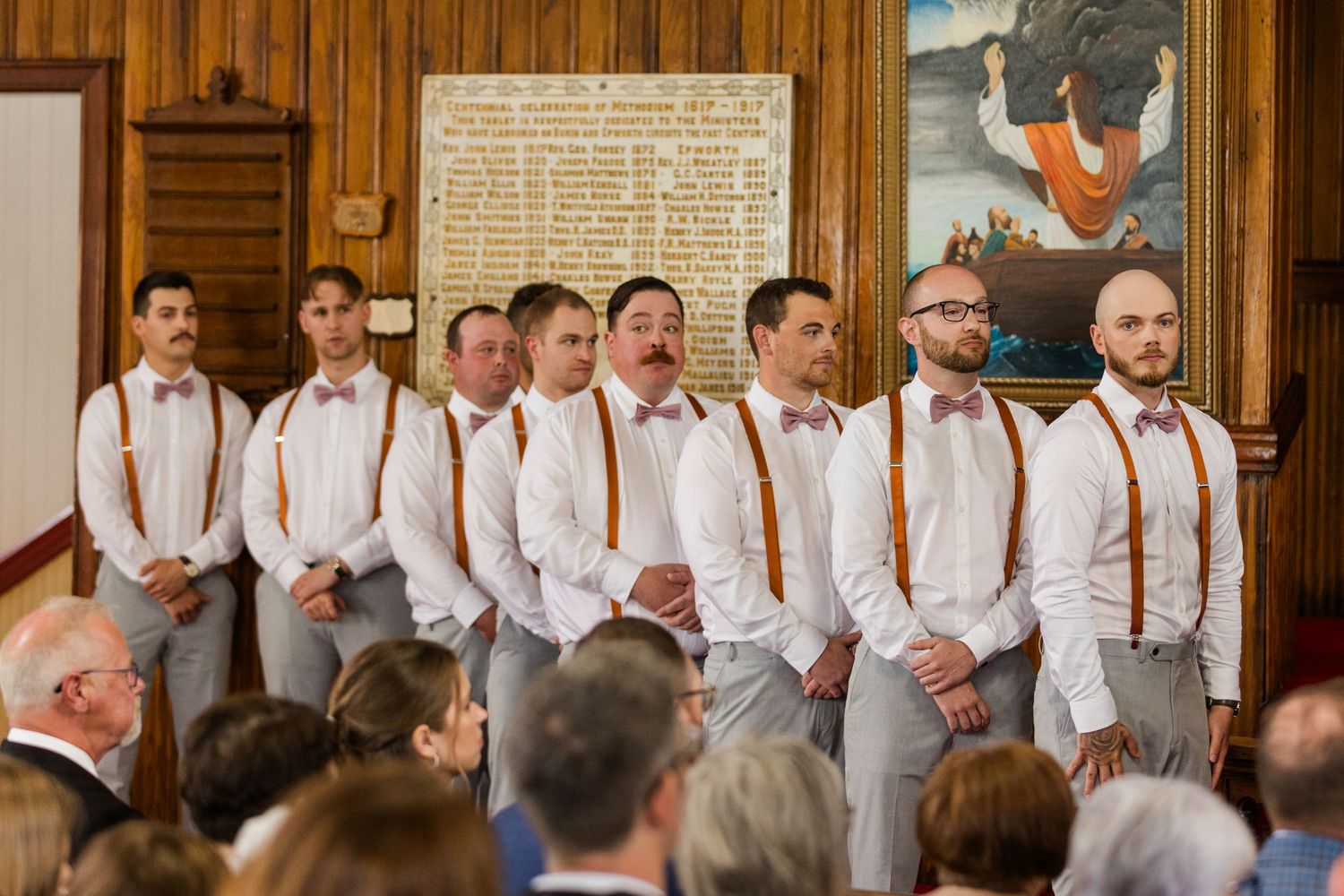 Wedding party members wearing white shirts and orange suspenders stand in a row at a formal ceremony venue.