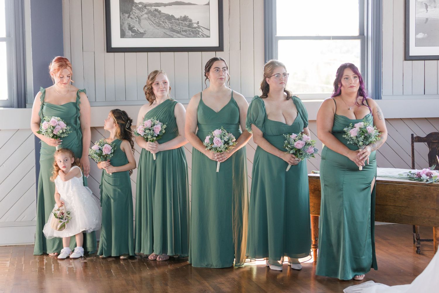 Wedding party in matching green dresses pose together in a rustic barn setting with white bouquets.