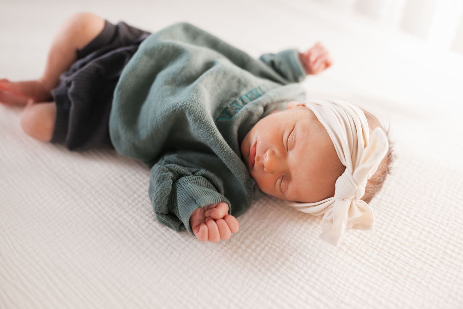 Sleeping newborn baby wearing teal outfit lying on white textured blanket.