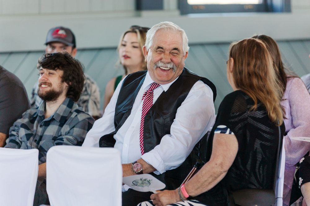 Audience members sit attentively during an indoor event with someone in a white shirt and red tie smiling in focus.