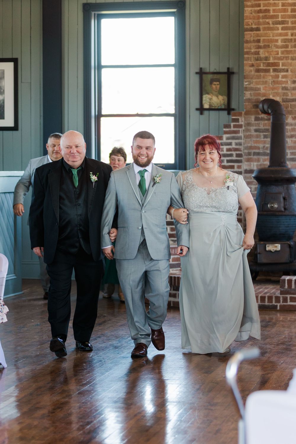 Wedding party members in grey suits and light colored dresses dance in a line on a hardwood floor at a rustic venue.