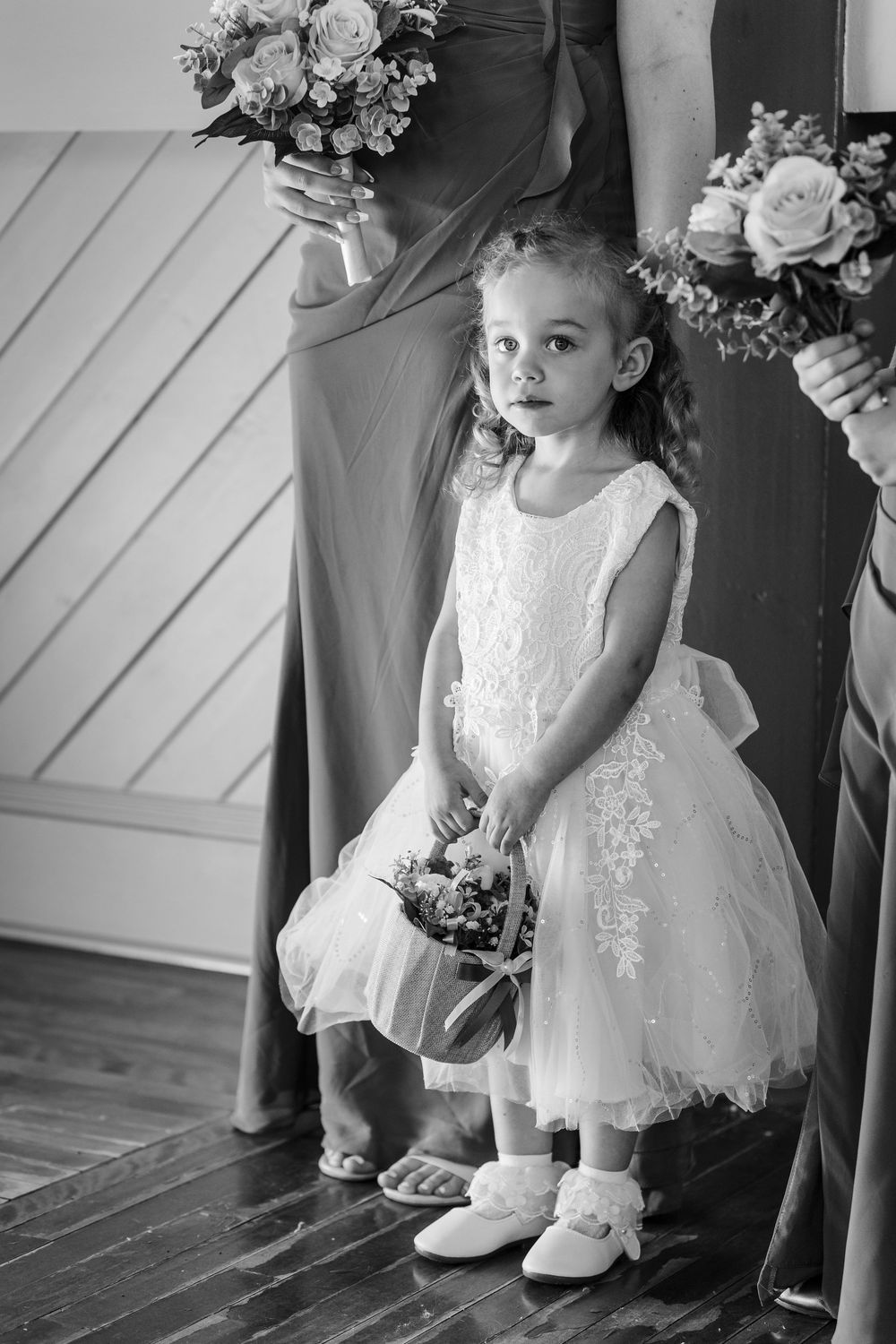 A flower girl in a white lace dress and shoes sits on a wooden floor at a wedding with bouquets visible nearby.